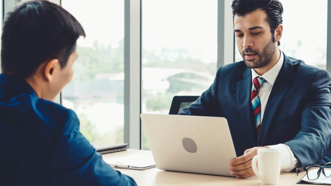Bold Leadership Path Solution Page Two men in business attire discussing over a laptop in a bright office setting.