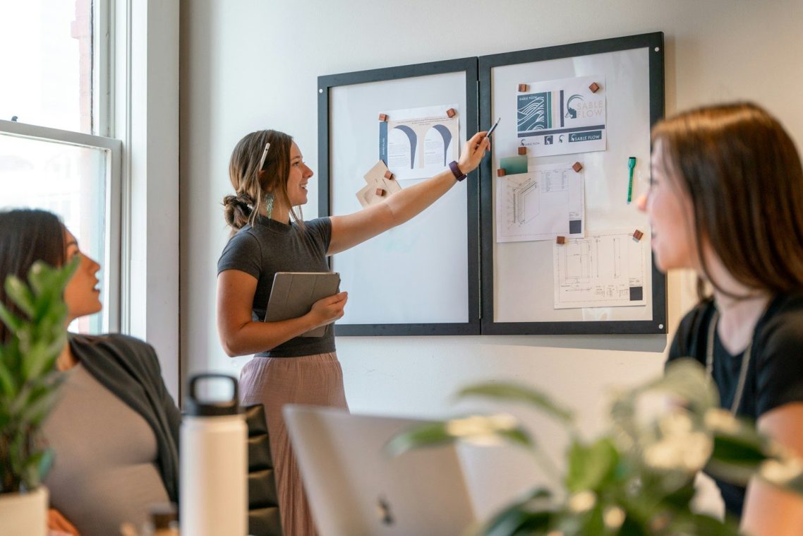 Bold Leadership Path The Method Page A woman presents ideas on a board to two colleagues in a modern office setting.