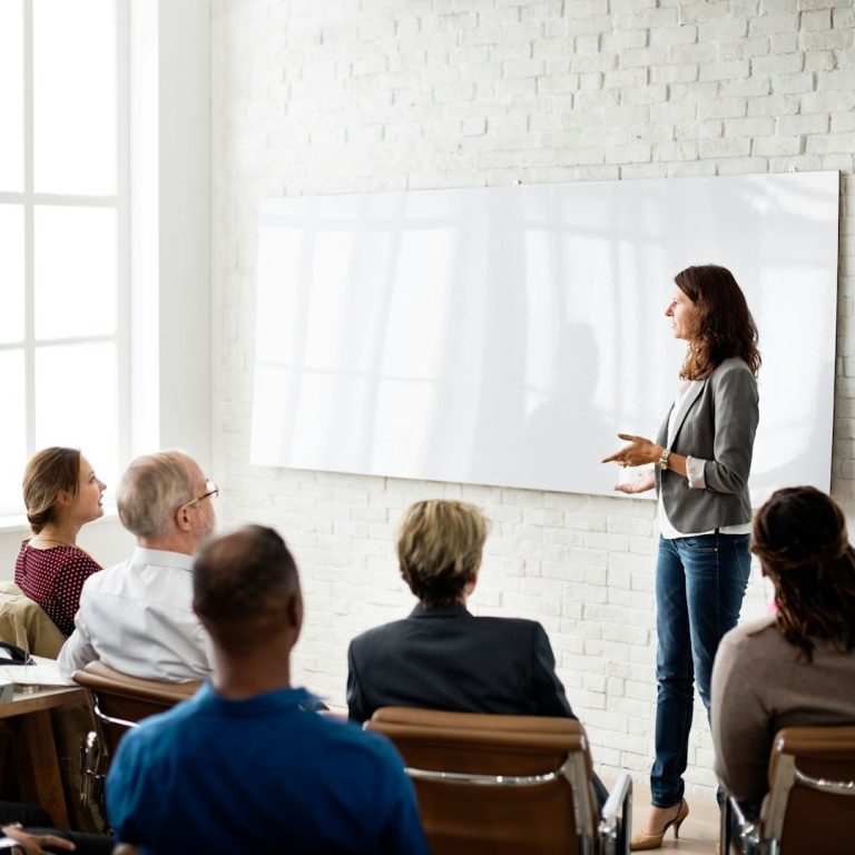 Bold Leadership Path logo on subscribe page” A woman presents to a group during a meeting in an office setting.